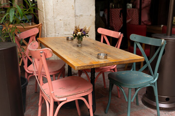 Rustic wooden table surrounded by colorful chairs in an outdoor cafe setting, adorned with a small vase of flowers, creating a cozy and inviting atmosphere for dining and relaxation