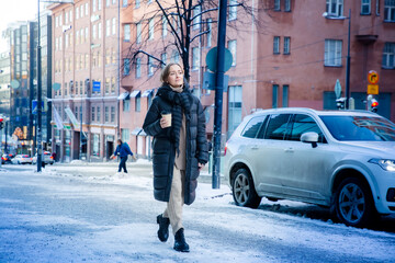 Middle aged woman walking along snowy city street with coffee cup, cozy winter urban lifestyle,...