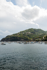 Scenic view of the green hills, white houses, and coastal architecture of Ischia island, Italy, seen from the Aragonese Castle. Boats and yachts float on the sea below, under dramatic weather.
