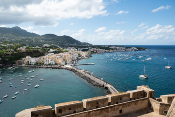 Scenic view of the green hills, white houses, and coastal architecture of Ischia island, Italy, seen from the Aragonese Castle. Boats and yachts float on the sea below, under dramatic weather.