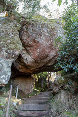 Stone steps leading beneath a massive moss-covered boulder at Hanging Rock, Macedon Ranges, Victoria, Australia. Scenic walking trail through the steeped in mystery, ancient volcanic rock formation.