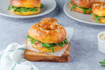 Three freshly baked bagels filled with greens and ham, on a wooden board and plates. Bagel breakfast sandwiches a zesty unique twist.