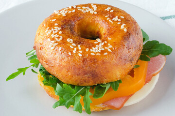 A delicious sesame bagel sandwich filled with ham, cheese, and fresh greens sits on a light plate. The meal is prepared on a textured surface, highlighting its freshness and color. 