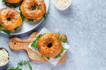 A selection of golden-brown bagels topped with sesame seeds sits on a wooden board. Fresh greens peek out from inside one bagel, complemented by a creamy spread nearby.