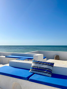 Enjoy the stunning scenery from a cozy rooftop in Djerba, Tunisia. The whitewashed terrace features blue seating, offering a perfect view of the sea and nearby palm trees.