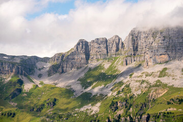 Fototapeta premium Dramatic rocky peaks of the Swiss Alps partly covered by clouds, with green alpine meadows and slopes below