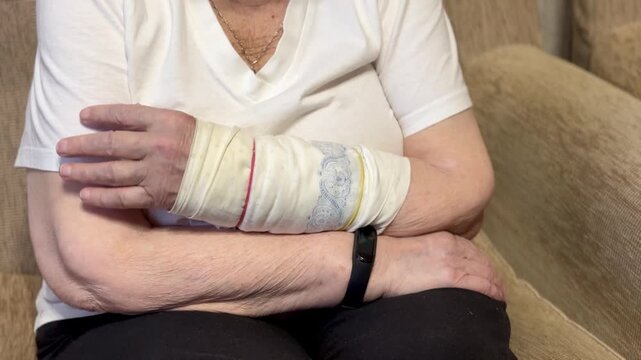 The black smart band with display touchpad. Portrait of an elderly grandmother with broken arm width cast for the arm sitting in armchair looks at the wristband of pulse monitor indoors