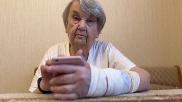 Elderly woman with broken arm width cast for the arm sitting at beige couch holding smartphone in her hand looking for important information using rose smartphone