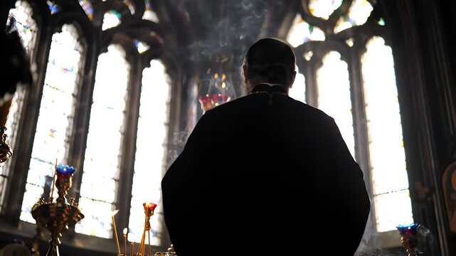 Silhouette of Priest in Dark Robes Standing in Church with Colorful Stained Glass Windows and Golden Candelabras