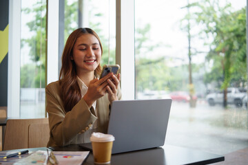 Obraz premium Young female entrepreneur checking mobile messages during remote work session at cafe with laptop and charts.