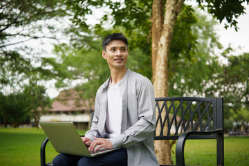 Fototapeta premium Young Asian man smiling while working on a laptop outdoors in a green park.