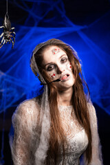 Portrait of creepy woman with horrible scary Halloween zombie makeup in white dress using wireless headset standing on blue background surrounded spiderweb. Devil Obsessed person. Horror