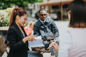 Multiracial business professionals engage in conversation outdoors, reviewing documents and sharing ideas during a casual meeting on a lively day.