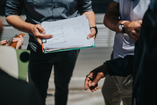 People collaborates over financial reports in an informal outdoor business meeting. The group examines data and graphs while engaging in a productive discussion, emphasizing teamwork and communication