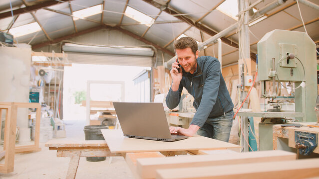 Carpenter Working In Woodwork Workshop Talking On Mobile Phone And Using Laptop