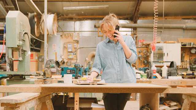 Female Carpenter Working In Woodwork Workshop Talking On Mobile Phone With Laptop Checking Order - Powered by Adobe
