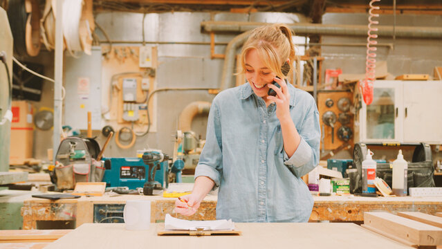 Female Carpenter Working In Woodwork Workshop Talking On Mobile Phone With Laptop Checking Order