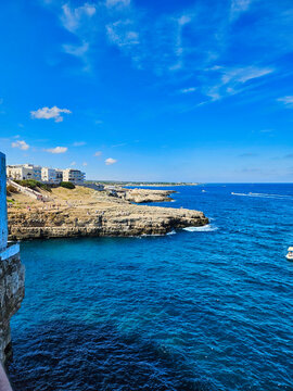 View from a rocky beach to a port with cranes and mountains in the background. White lighthouse on the island with blue sea, big rocks and green trees. La costa con il mare verde smeraldo.