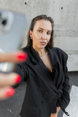 Confident woman in oversized black blazer taking a selfie with smartphone outdoors, representing modern fashion, technology, and urban lifestyle.