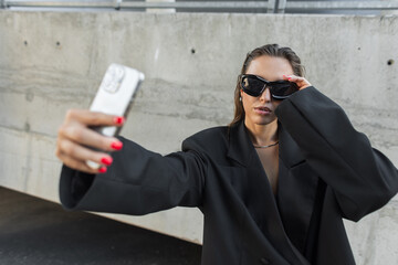 Stylish woman in oversized black blazer and sunglasses posing for a selfie with smartphone outdoors, showcasing urban fashion and modern confidence.