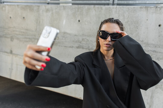 Stylish woman in oversized black blazer and sunglasses posing for a selfie with smartphone outdoors, showcasing urban fashion and modern confidence.