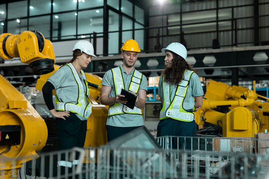 Team of engineers and robot arm factory worker wearing safety helmets and reflective vests using digital tablet while discussing project in modern industrial workplace. Concept of teamwork.