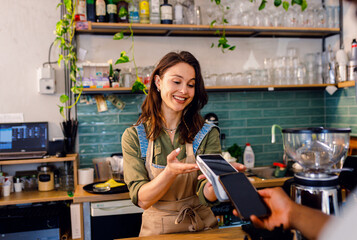 Female barista charges the customer for takeaway coffee in cozy modern coffee shop.