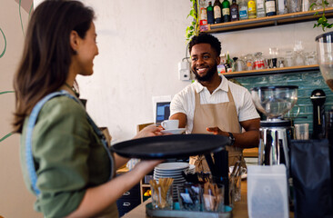 Male barista working in a coffee shop at the counter and gives a coffee to a female waiter to serve to the customer.