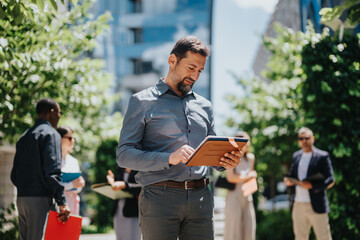 A businessman using a tablet in an outdoor setting, surrounded by colleagues in a leafy urban area, symbolizing teamwork and innovative collaboration.
