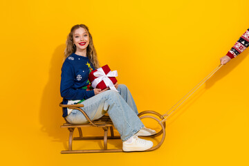 Happy young woman with Christmas gift sitting on sled and enjoying festive atmosphere against yellow background