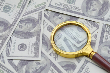 A golden magnifying glass rests on a pile of one-hundred-dollar bills, symbolizing a close...