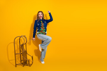 Fototapeta premium Festive Celebration with Young Woman in Christmas Sweater Posing Playfully with Sleigh Against Bright Yellow Background