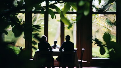 Silhouetted figures gather around a table indoors framed by lush green foliage and large windows overlooking a natural landscape