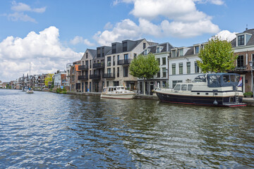 Boats moored along the Oude Rijn river in Alphen aan den Rijn, with modern and classic Dutch buildings lining the waterfront under a blue sky.