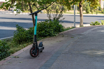 Green electric scooter standing on sidewalk near road and trees, urban eco transport option for modern city lifestyle.