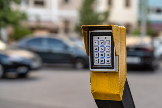 Electronic keypad device mounted on yellow post in outdoor parking area, secure access control technology with blurred cars in background.