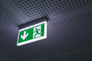 Illuminated green exit sign with downward arrow and running figure, mounted on perforated ceiling. Safety, emergency, direction.