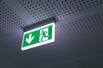 Green illuminated emergency exit sign with arrow pointing down, mounted on ceiling. Safety, escape.