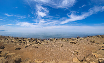 Wide angle shot of rocky shore with a calm sea under a bright blue sky. White clouds stretch across the horizon, creating a peaceful coastal landscape full of natural beauty and open space.