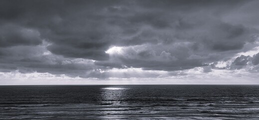 A dramatic seascape banner where sunlight pierces through heavy clouds, casting radiant beams onto a calm ocean. The interplay of light and shadow creates a powerful yet peaceful atmosphere