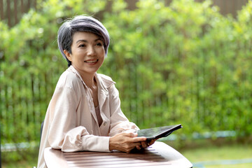 Asian adult woman happy using digital tablet sitting outdoor garden working business browsing internet connection mobile technology device relax lifestyle