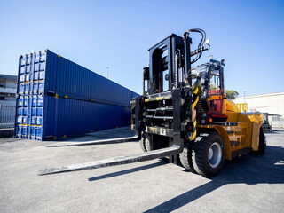 Forklift at a container terminal. A yellow and black heavy-duty forklift stands in the foreground on an industrial yard. Its long, massive forks are clearly visible, pointing toward the center