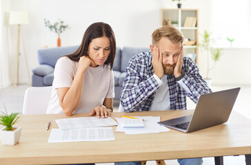 Fototapeta premium Sad focused young couple calculating bills in worry, discussing planning budget together, serious wife and husband looking at laptop screen, using online banking services and calculator, poor finances