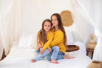 Happy family time concept. Sisters sit in bed at the morning and smiling in camera. Smiling teen girl embracing little sister siblings playing relaxing on the bed. Child friendship together concept. 
