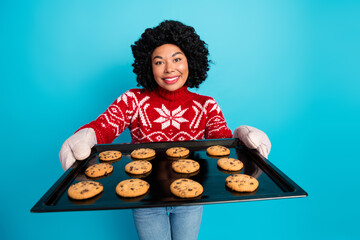 Smiling woman in festive sweater holding a baking tray of cookies on a vibrant blue seasonal background