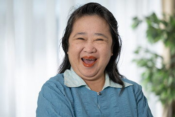 Asian Happy middle aged woman smiling with crossed arms in casual blue shirt indoors with blurred...