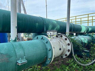 An industrial pipe network is shown in the image, featuring a flow meter connected with flanges. The pipes are painted green.Industrial infrastructure featuring a large, weathered pipe with a control 