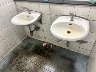 This image shows two dirty sinks in a neglected public restroom, highlighting poor hygiene and unsanitary conditions. The cracked tiles and grimy floor suggest a lack of maintenance and sanitation.
