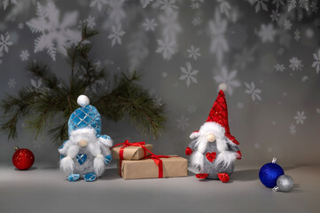 

Two festive gnome figurines decorated with red and blue attire stand beside Christmas gifts wrapped with red ribbon. Ornaments add a cheerful holiday vibe to this winter scene.