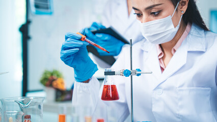 A gloved scientist carefully drips red solution into a flask, with red test tubes in the...
